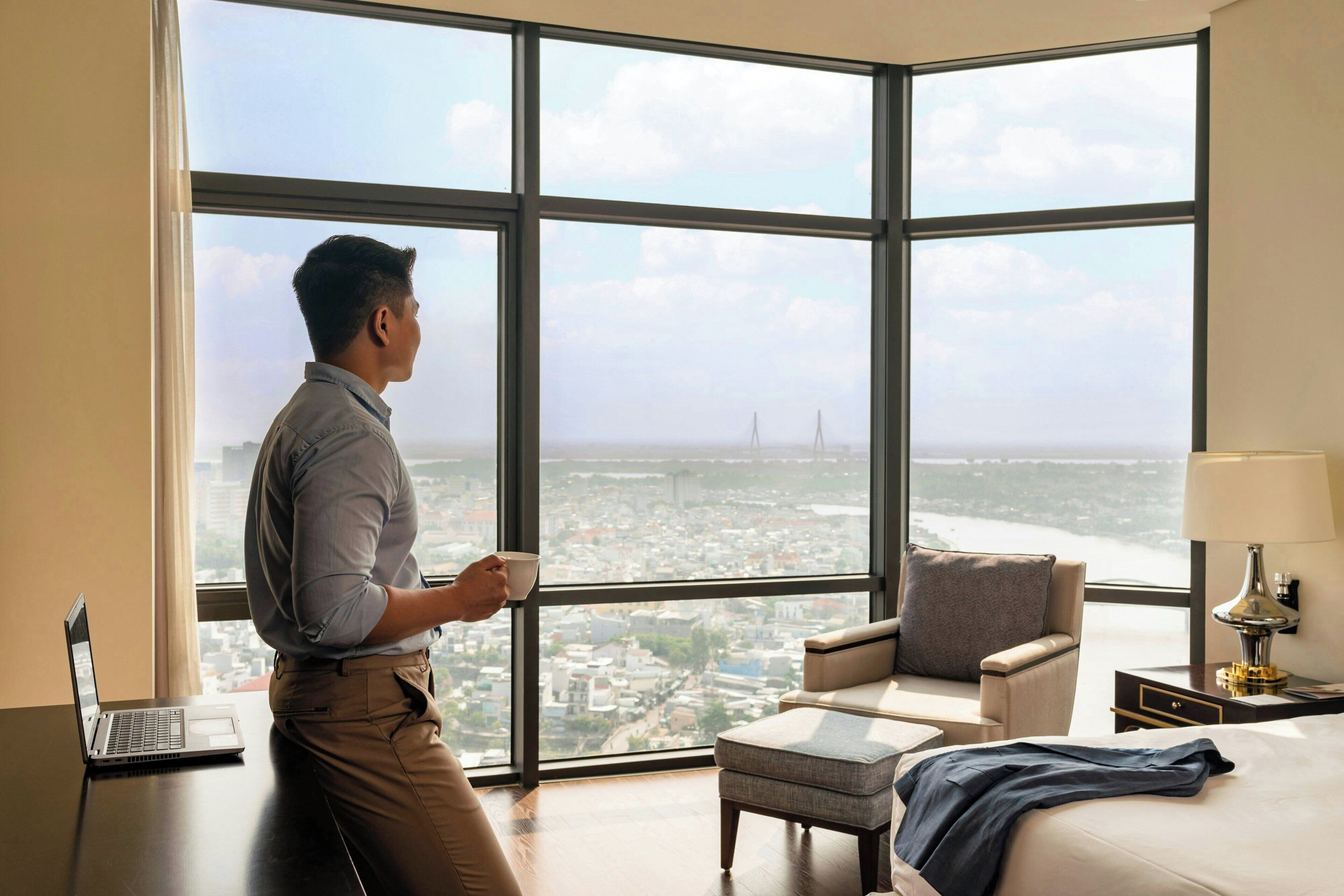 A hotel room with a large window, a laptop on a desk, a bed, a chair, and a nightstand with a lamp.