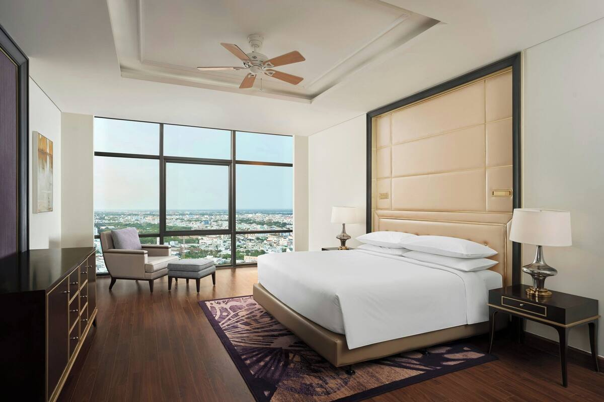Amplia habitación de hotel con una cama grande, un área de descanso con una silla, una mesita de noche con lámparas y vistas a la ciudad a través de amplios ventanales.