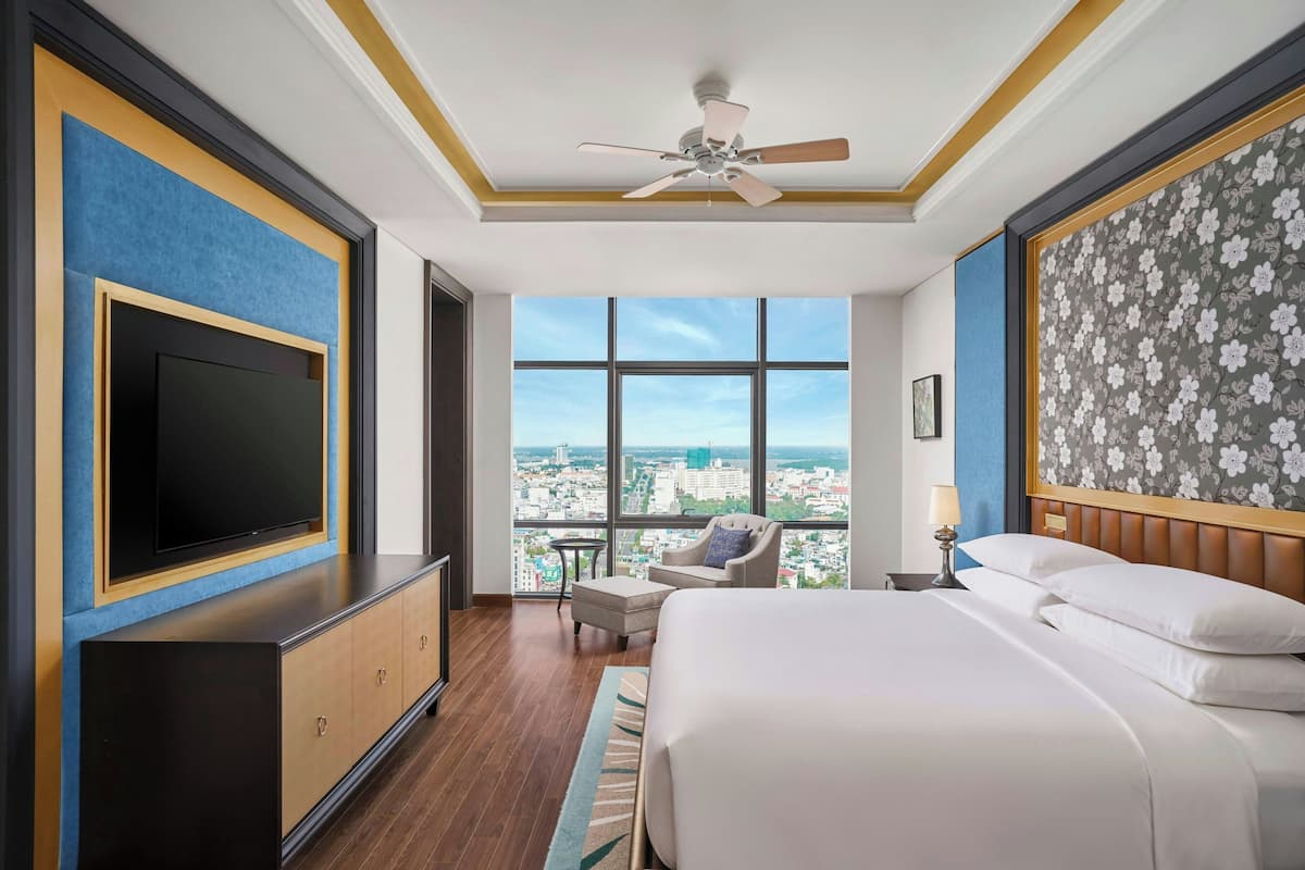Habitación de hotel con una cama grande, un televisor de pantalla plana, un ventilador de techo y vistas a la ciudad a través de amplias ventanas.