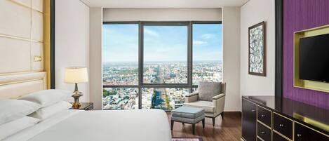 A hotel room with a large bed, a desk, a chair, a TV, and a view of the city.