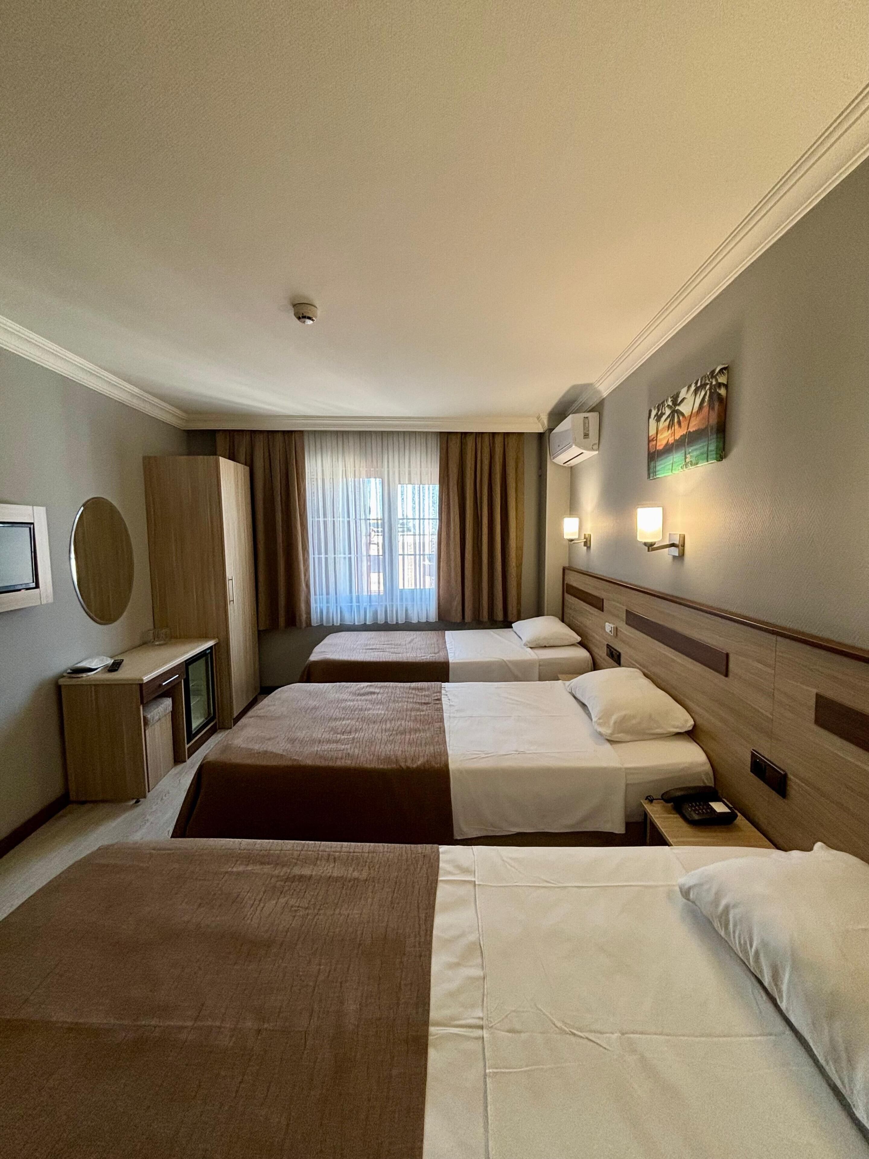 A hotel room with two beds, a desk, a mirror, a wardrobe, and a painting on the wall.