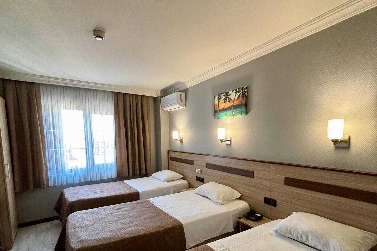 A hotel room with three beds, a large window with curtains, an air conditioning unit, and wall-mounted lights.