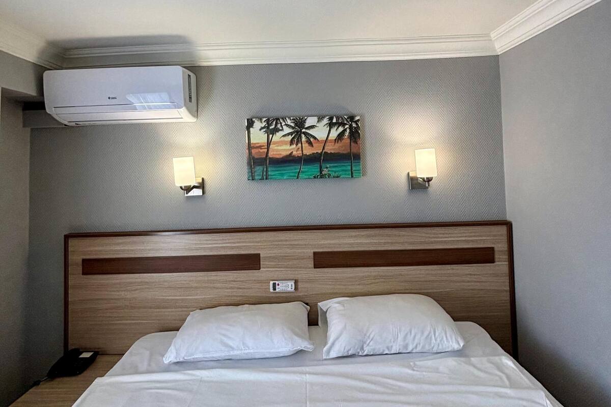 A hotel room with a bed, an air conditioning unit, a nightstand, a wall-mounted lamp, and a painting of a beach scene.