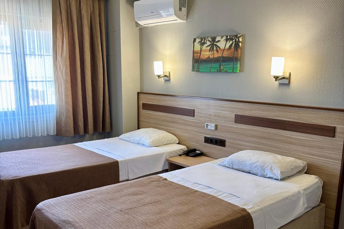 A hotel room with two beds, a ceiling fan, an air conditioning unit, a painting, and a wall-mounted light fixture.