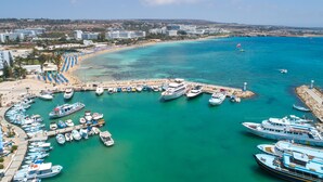 A marina with various boats docked, a beach with umbrellas, and buildings in the background. - Blue Harbour Boutique (Ayia Napa)