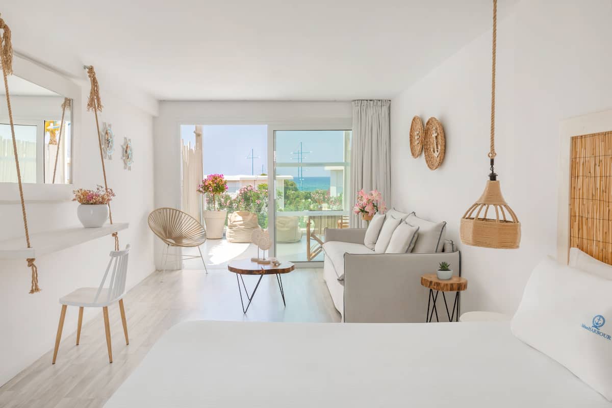 A bright, modern living room with a sofa, chairs, a small table, and a view of the sea through large windows.