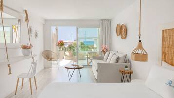 A bright, modern living room with a sofa, chairs, a small table, and a view of the sea through large windows.