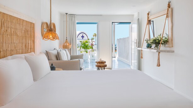 A modern bedroom with a large bed, a sofa, a small table, and a balcony with a view of a medical building. - Blue Harbour Boutique (Ayia Napa)