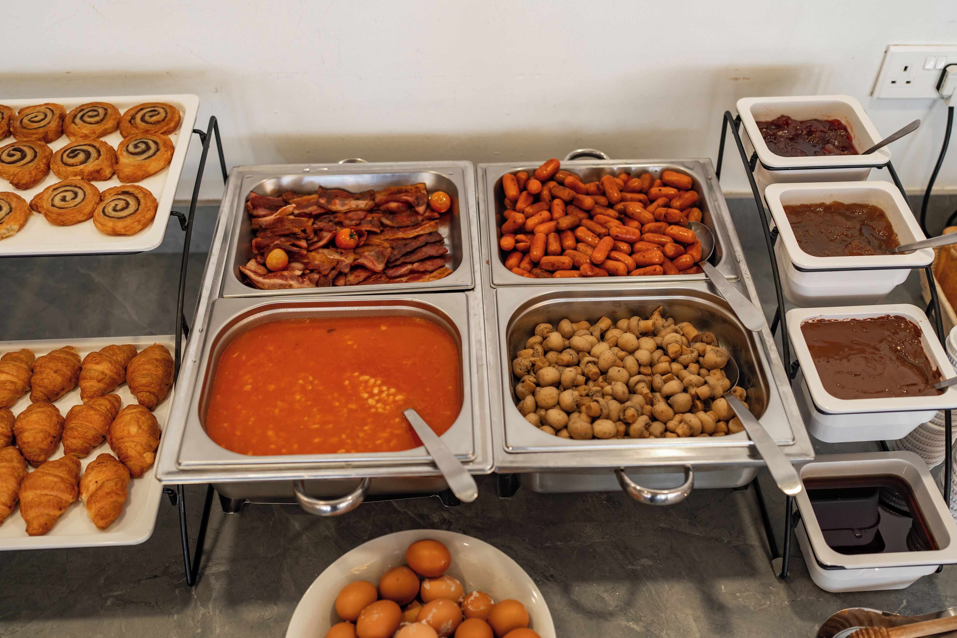 A buffet with various food items including pastries, bacon, sausages, and condiments.