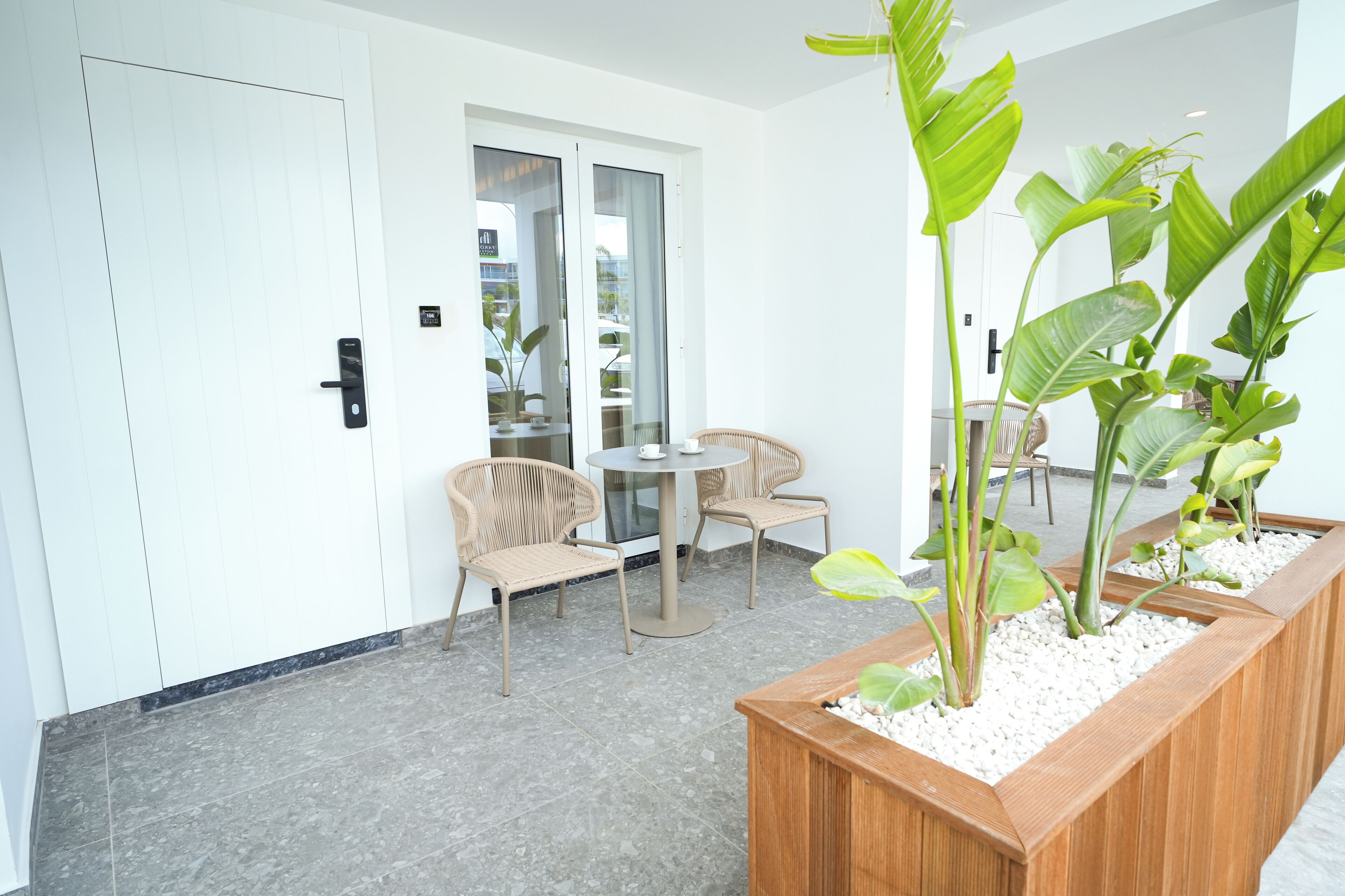 Une chambre d’hôtel moderne avec un banc en bois, deux fauteuils en osier, une table ronde et un grand pot contenant des plantes vertes.