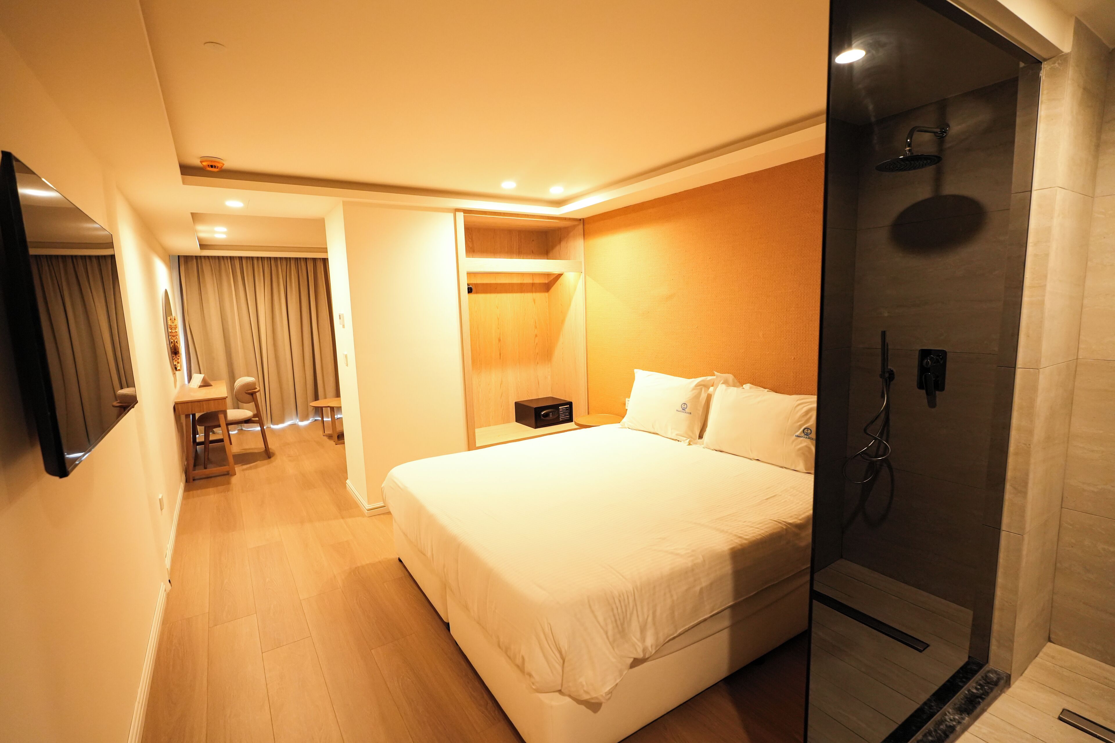 A hotel room with a large bed, a bedside table, a desk with a chair, a shower area, and a mirror.