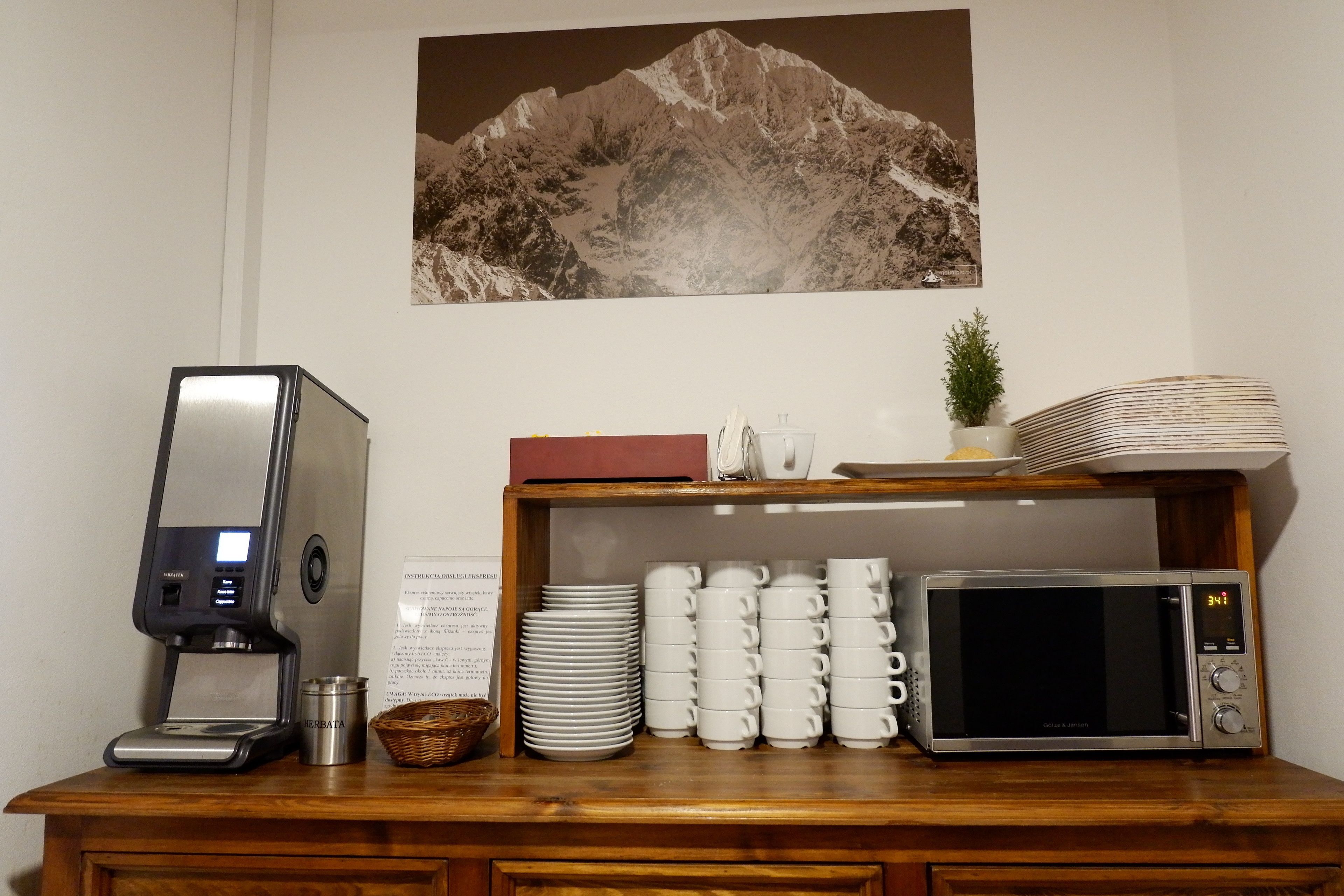 A coffee station with a coffee maker, a microwave, and stacked white cups on a wooden counter.