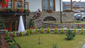 Une maison en bois à deux étages, dotée d’une façade en pierre, d’un balcon avec des balustrades en bois et d’un jardin où poussent diverses plantes et fleurs.