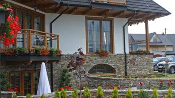 A two-story wooden house with a balcony, stone facade, and a garden with various plants.