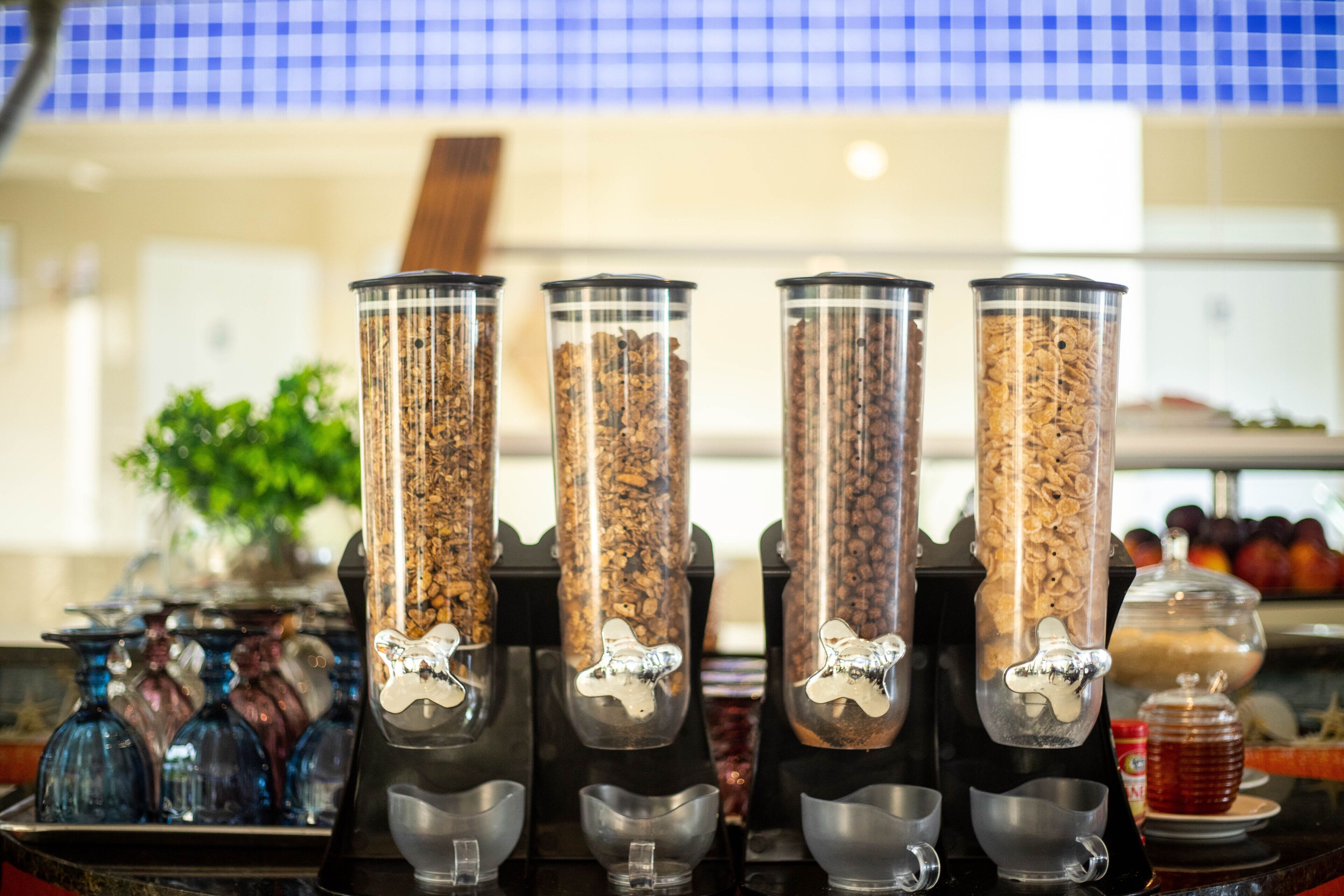 Cuatro dispensadores de café con un asa metálico, una bandeja con botellas de vidrio azul y una exhibición de frutas al fondo.