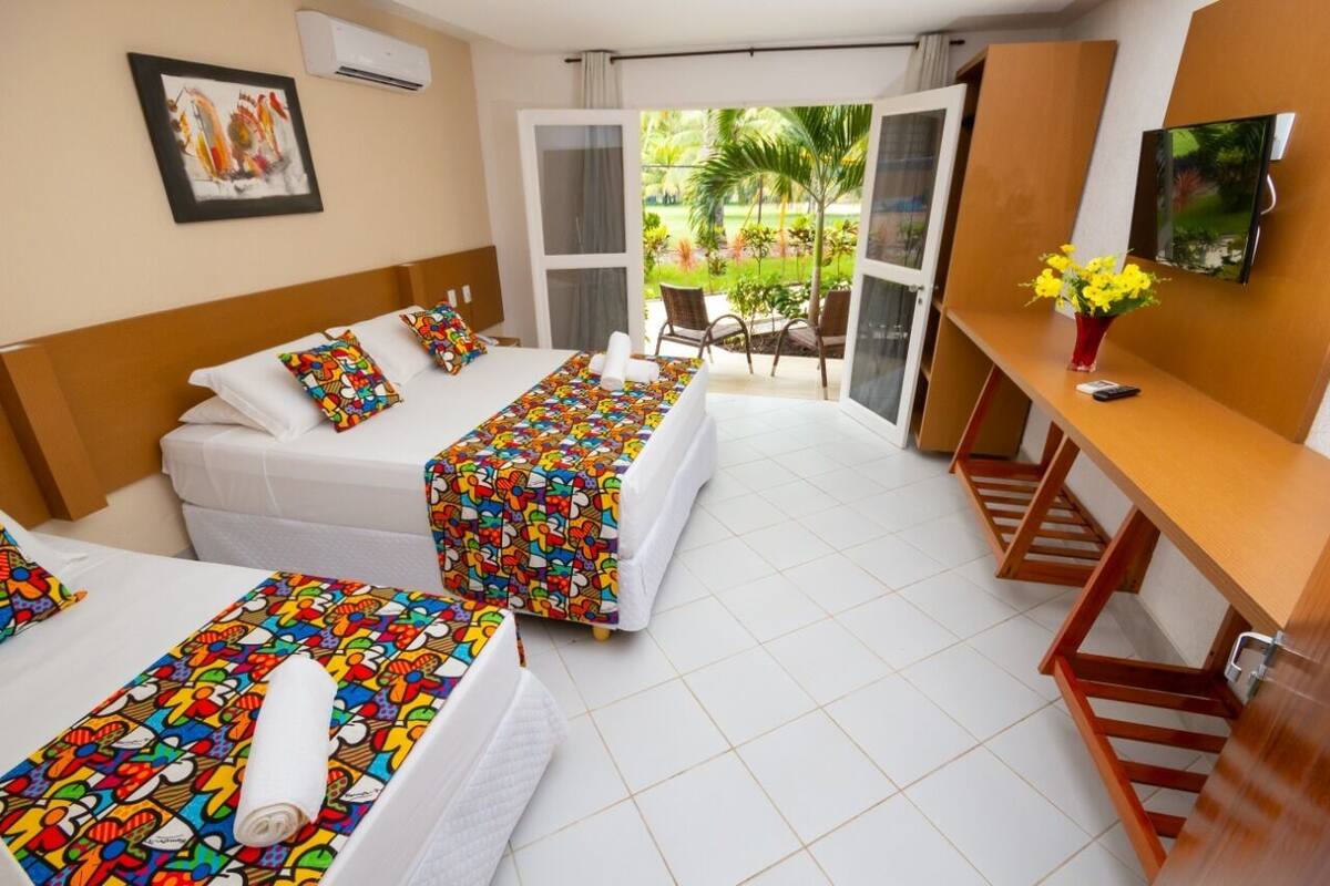 A hotel room with two beds, a TV, a desk, and a balcony with a view of the garden.
