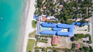 An aerial view of a resort with multiple blue-roofed buildings, a beach, and palm trees. - Crôa Mares Hotel (Maragogi)
