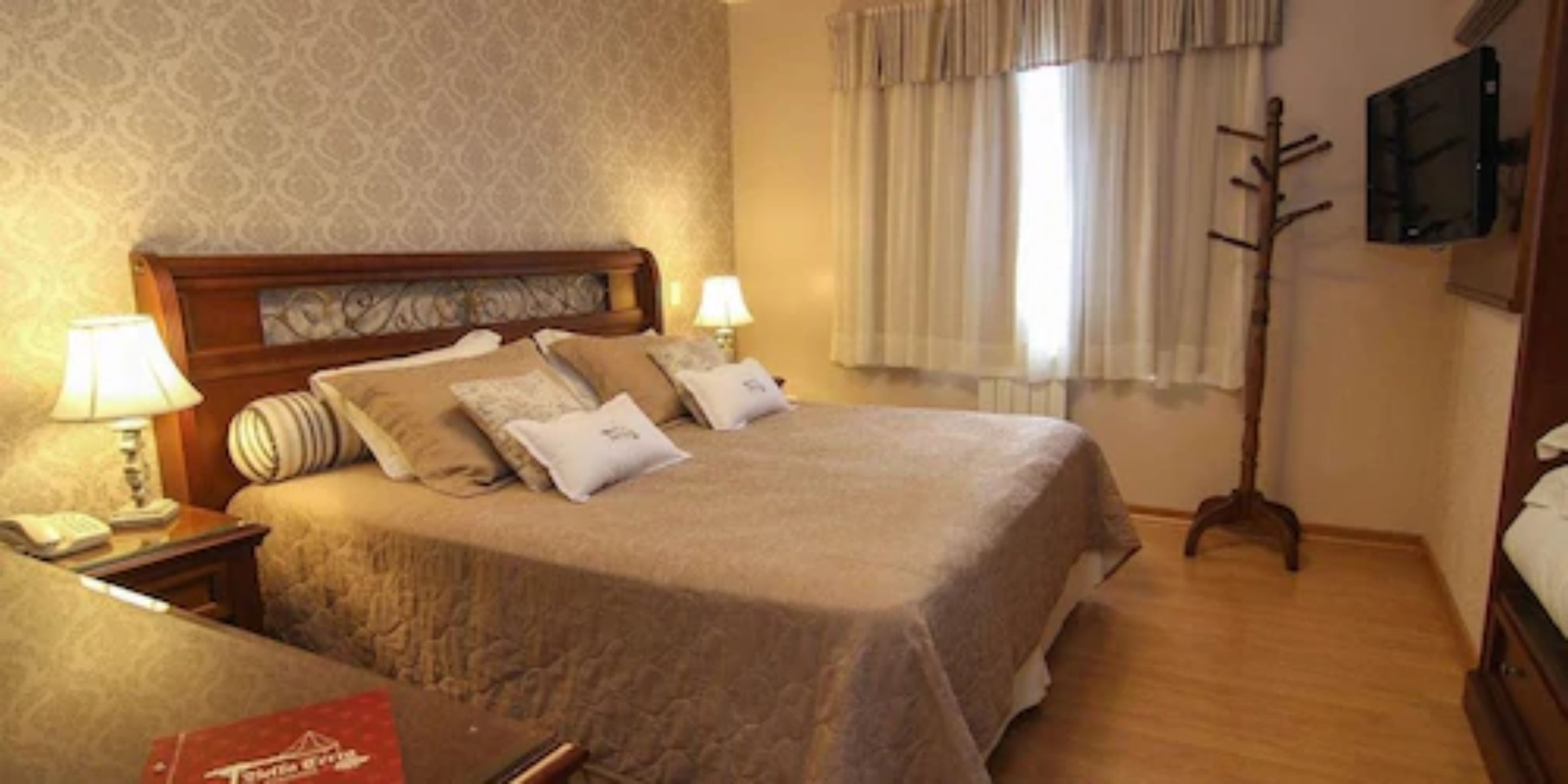 Une chambre d’hôtel avec un grand lit, deux lampes de chevet, une table de nuit en bois, un livre rouge, un porte-manteau en bois et un téléviseur fixé au mur.