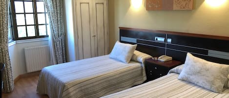 Quarto de hotel com duas camas, uma janela com cortinas, um radiador, um guarda-roupa e um candeeiro fixado na parede.