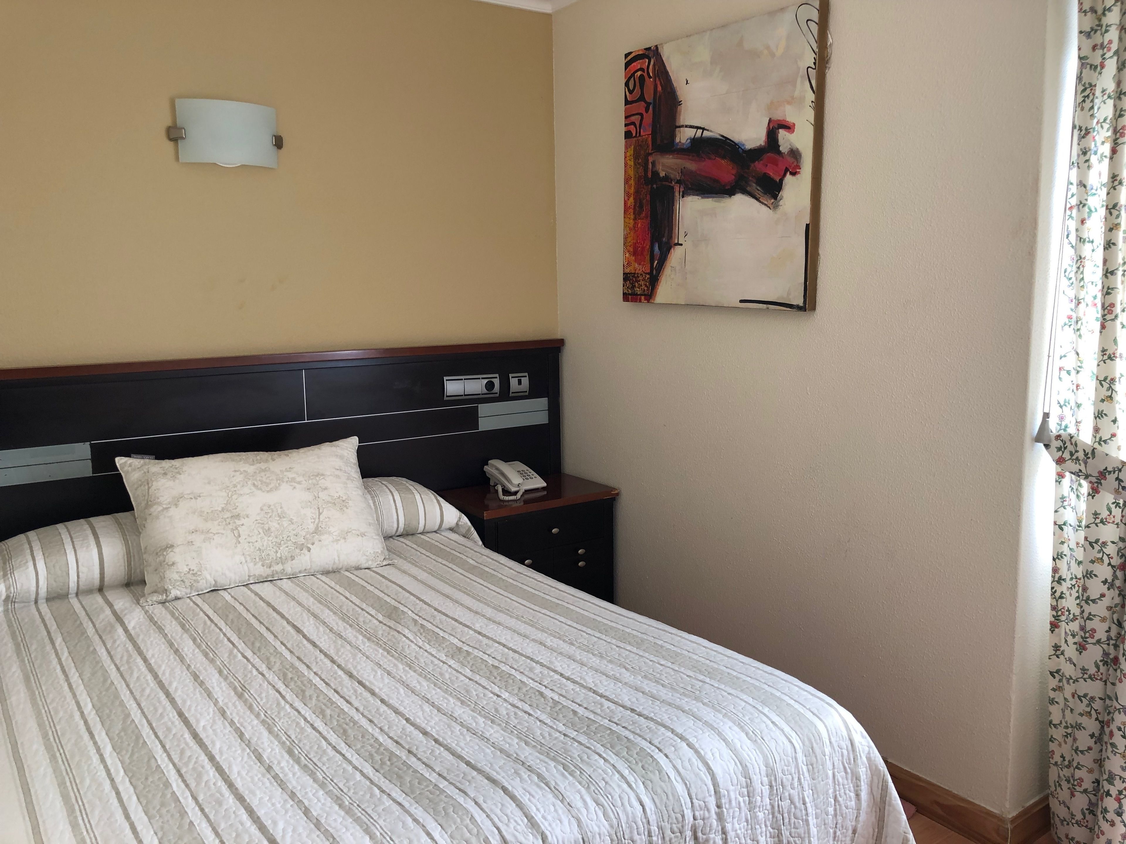 A hotel room with a bed, a nightstand, a telephone, a wall-mounted light, and a painting.