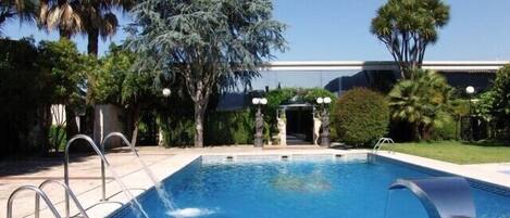 Una piscina con escaleras metálicas, rodeada de palmeras y un edificio al fondo.