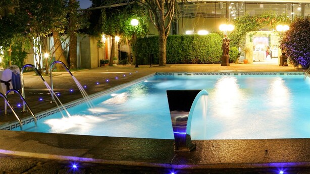 A swimming pool with a central water feature, surrounded by a well-lit patio area with lounge chairs and decorative lighting. - Hotel Sena (Caldas de Reis)