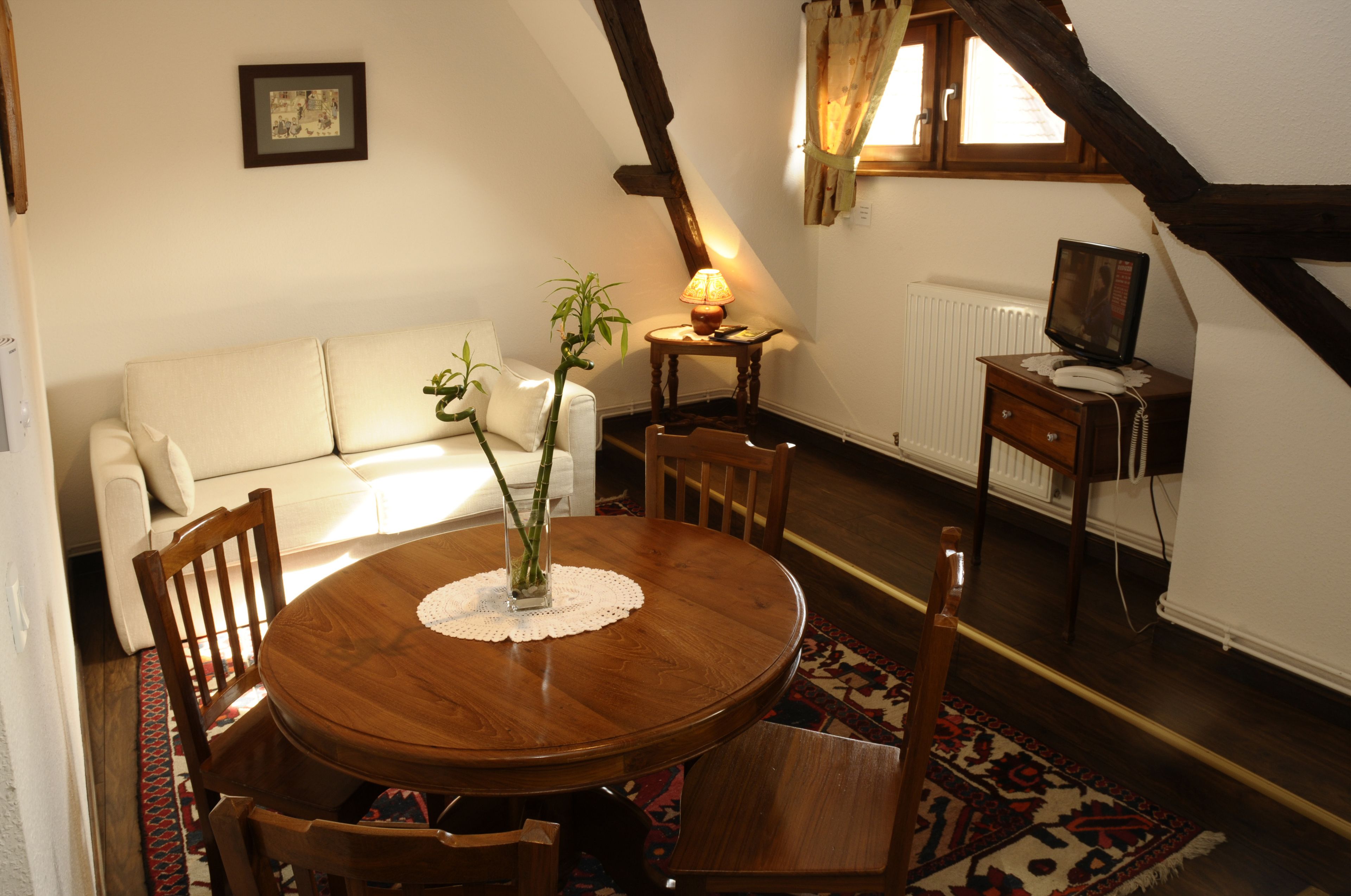 A cozy room with a wooden dining table, chairs, a white sofa, a small side table with a lamp, a radiator, and a television on a wooden stand.