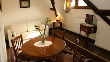 A cozy room with a wooden dining table, chairs, a white sofa, a small side table with a lamp, a radiator, and a television on a wooden stand.