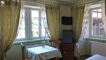 Una habitación con una cama, una mesa con un florero, un televisor sobre un mueble y cortinas.