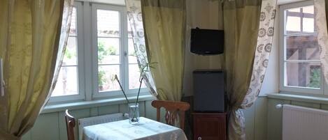 Una habitación con una cama, una mesa con un florero, un televisor sobre un mueble y cortinas.