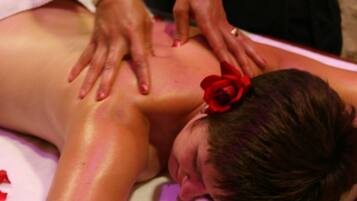 A massage therapy session in progress with a client lying face down on a massage table, a therapist's hands on the client's back, and a red rose accessory in the client's hair.