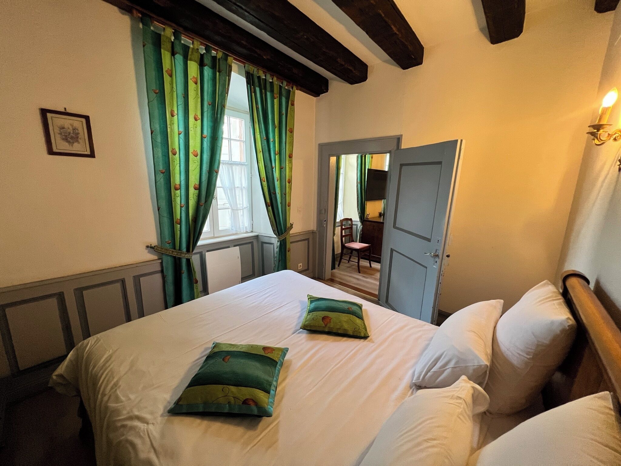 A hotel room with a bed, green curtains, a wooden ceiling, a door, and a small table with a chair.