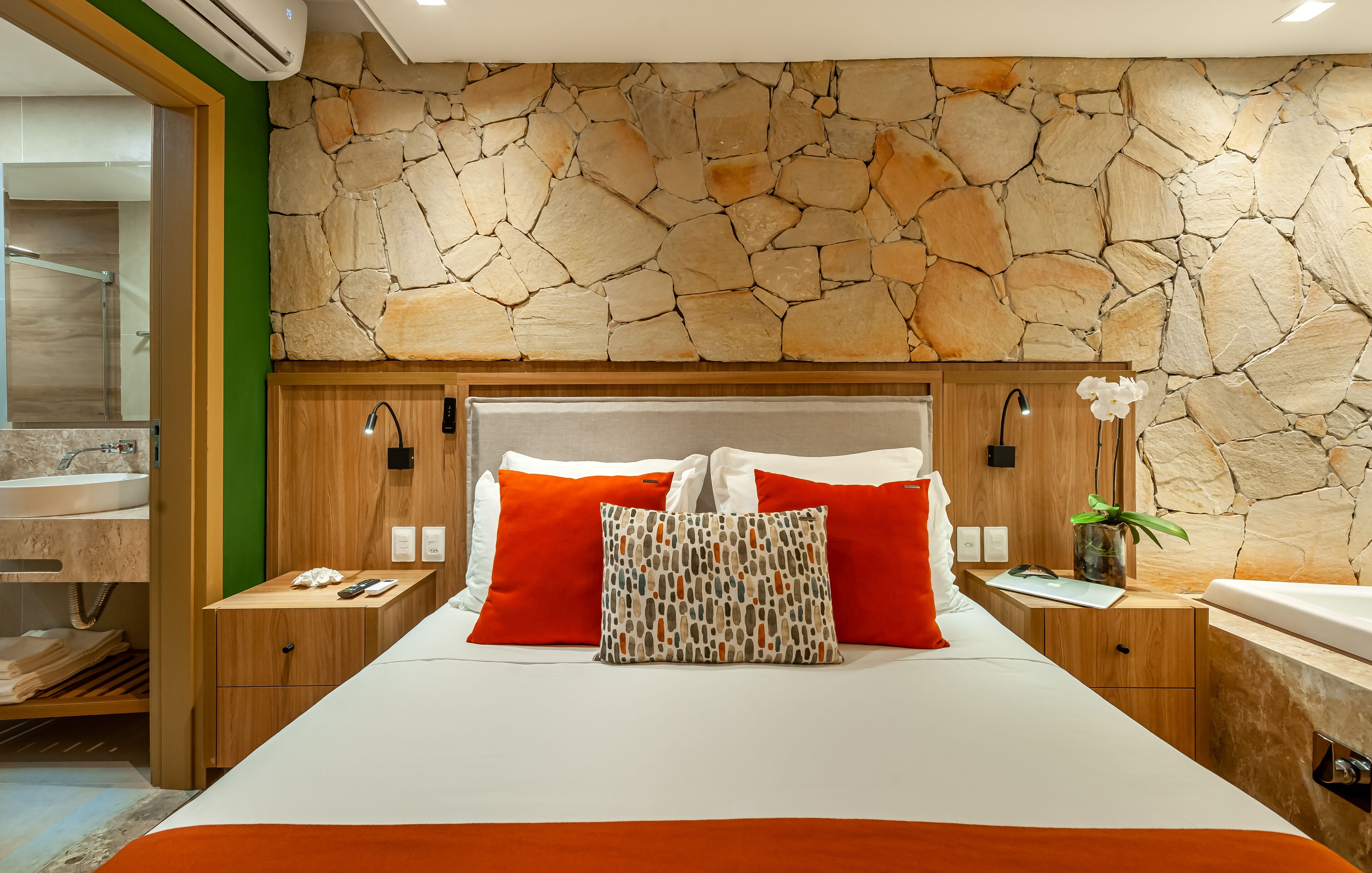 A modern hotel room with a stone wall, wooden headboard, and a large bed with red and white bedding.