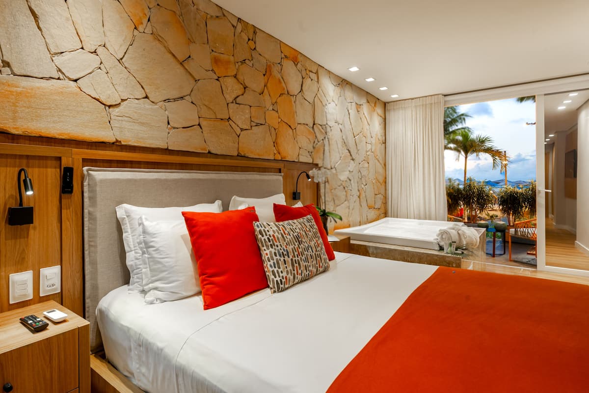 Habitación de hotel con pared de piedra, cama con ropa de cama blanca y roja, mesita de noche con teléfono y control remoto, y vista a la playa.