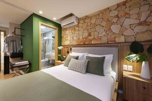 Habitación de hotel moderna con pared de piedra, cama con muchas almohadas, una prenda colgada y una maleta en el suelo.