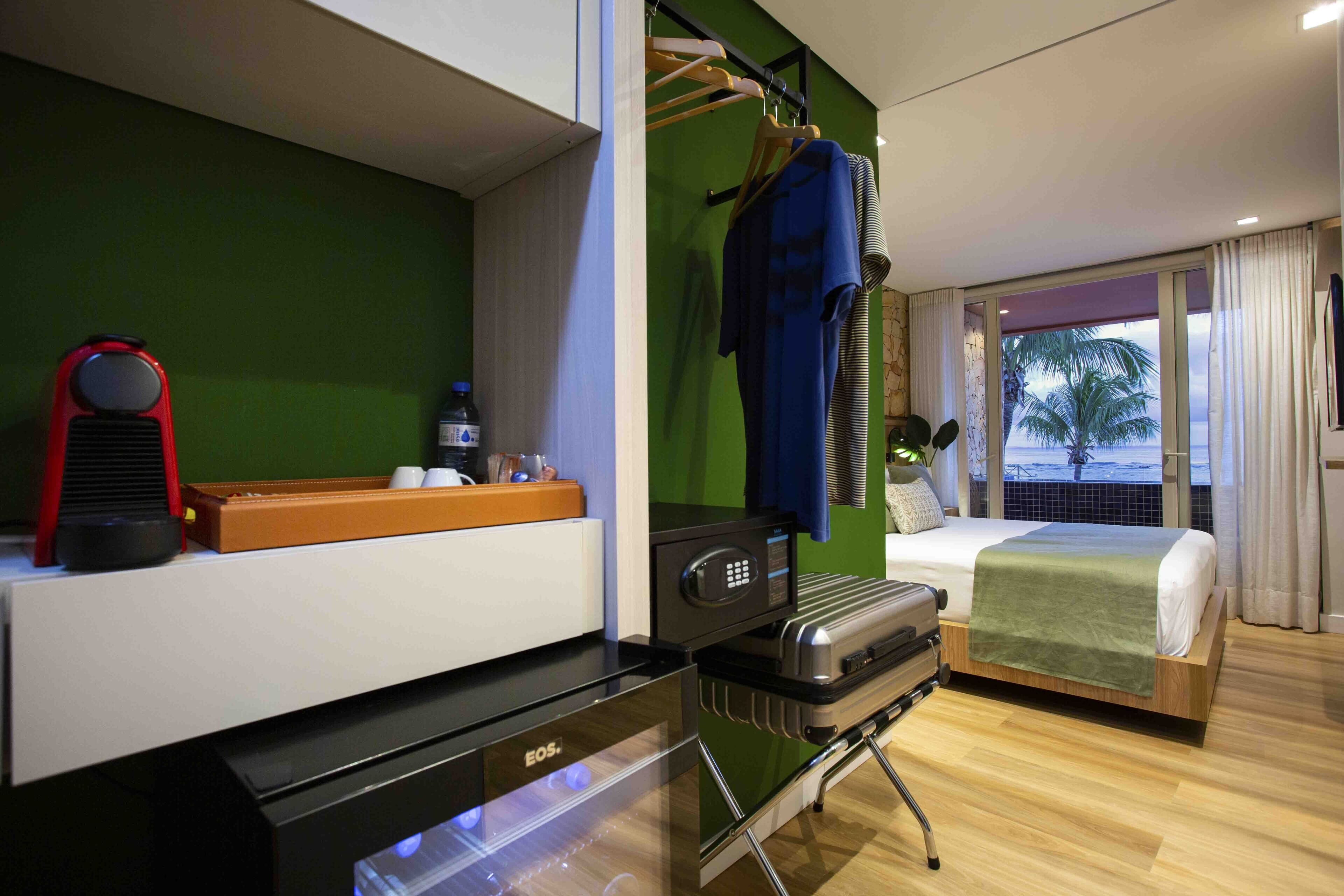 A modern hotel room with a bed, a suitcase, a microwave, a coffee maker, and a view of the beach through the window.
