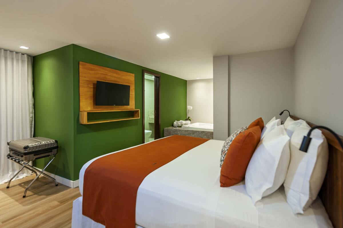 Una habitación de hotel con una cama, un televisor montado en un panel de madera, un baño con bañera y una maleta en el suelo.