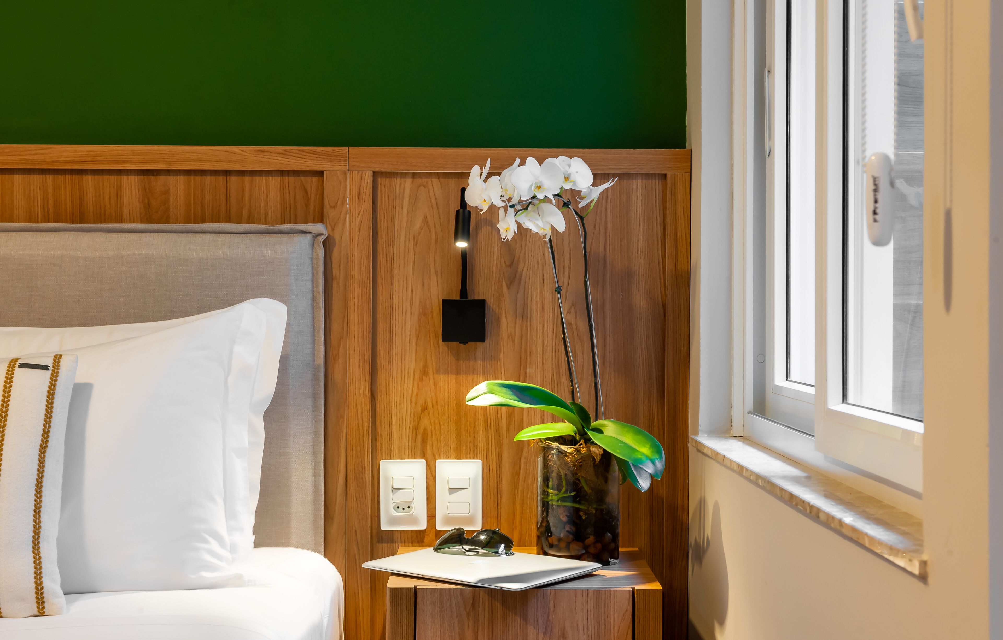 Een keurig opgemaakte hotelkamer met een bed, nachtkastje met een vaas orchideeën, en een raam met uitzicht.
