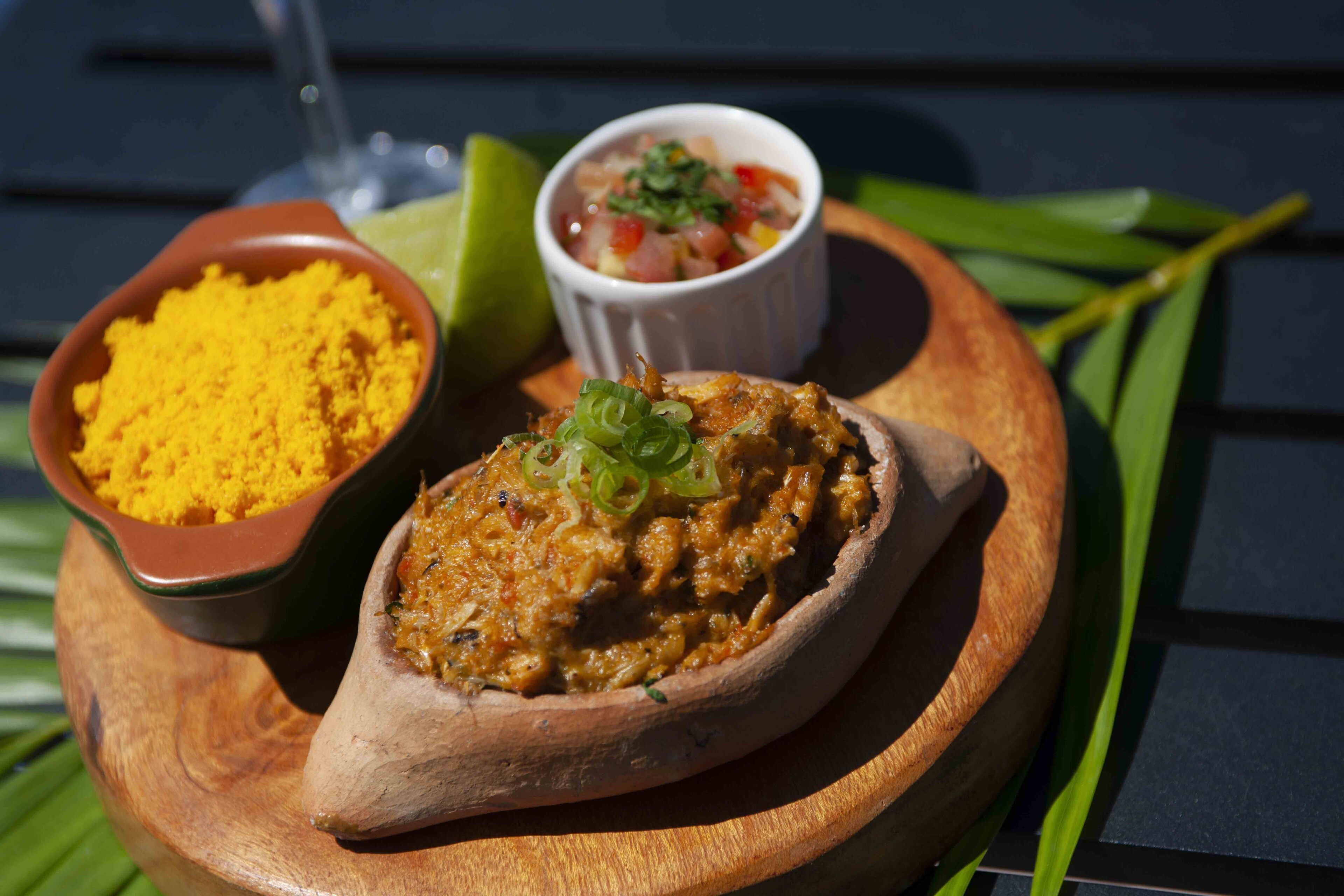 Uma tábua de madeira com um prato de tempero amarelo, uma tigela de salsa e um pimentão recheado.