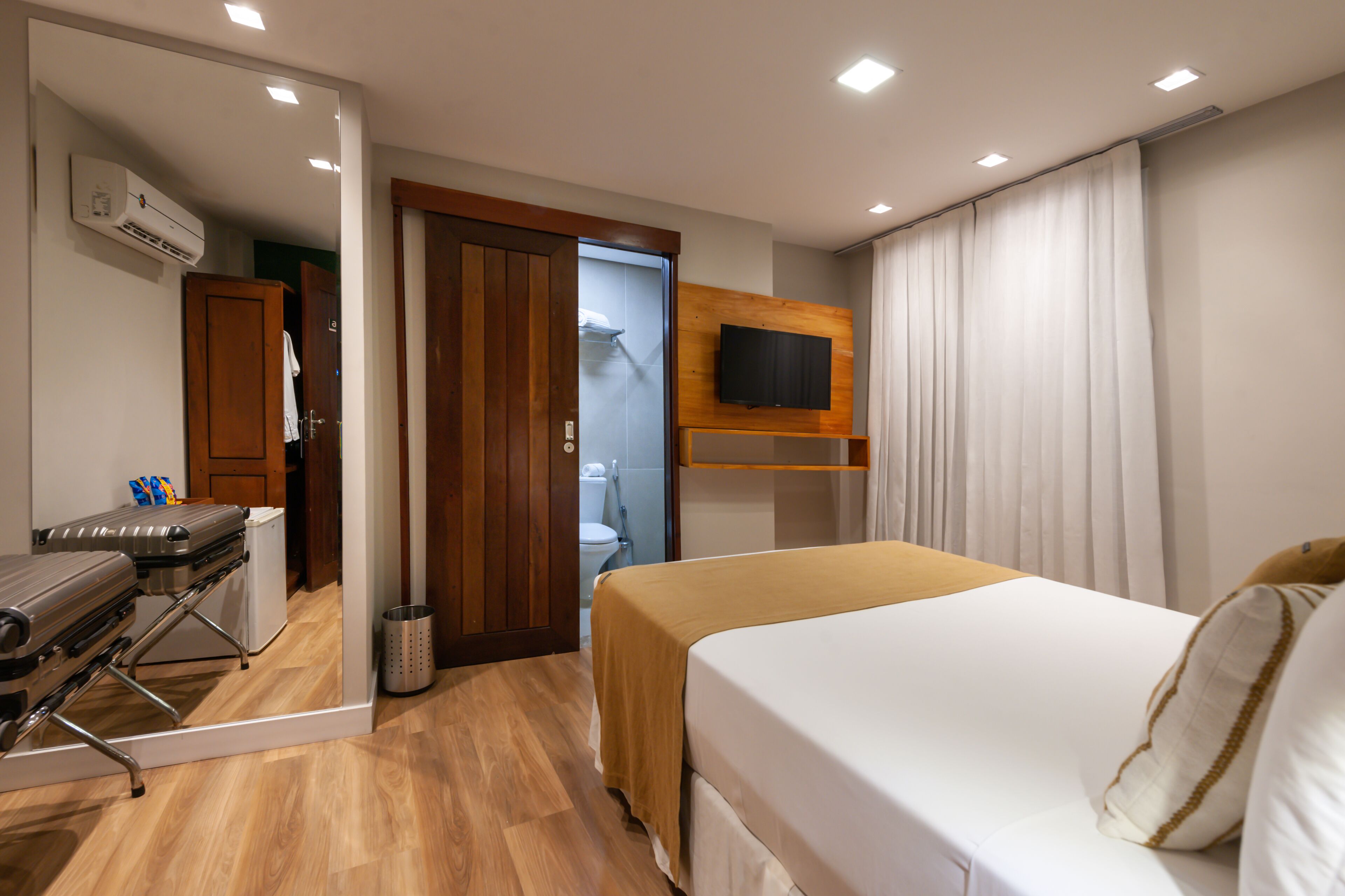 A hotel room with a bed, a TV mounted on the wall, a wooden floor, a mirror, and a suitcase.