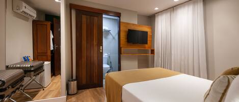 A hotel room with a bed, a TV mounted on the wall, a wooden floor, a mirror, and a suitcase.
