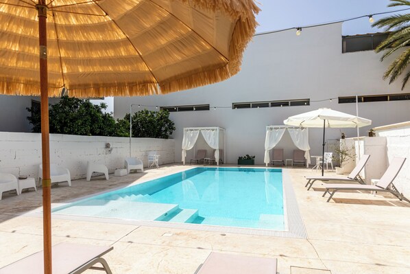 A swimming pool with lounge chairs and umbrellas, surrounded by a white building with windows and a wall of greenery. - La Gemma del Salento (Racale)