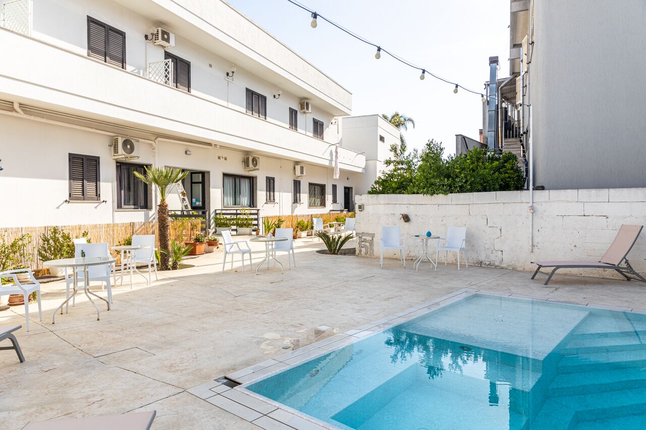 A pool area with a clear pool, white outdoor furniture, and a building with balconies and air conditioning units.