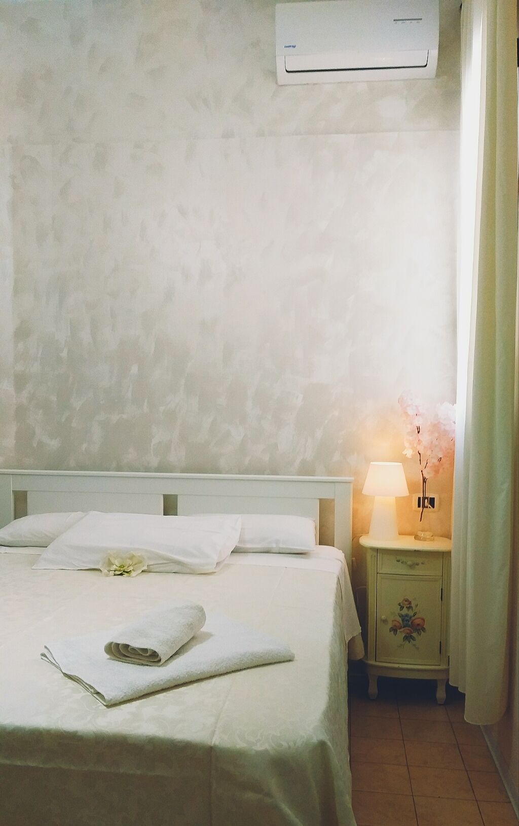 Un lit bien fait, avec du linge de lit blanc et une serviette pliée, une table de chevet avec une lampe et une commode à motifs floraux.