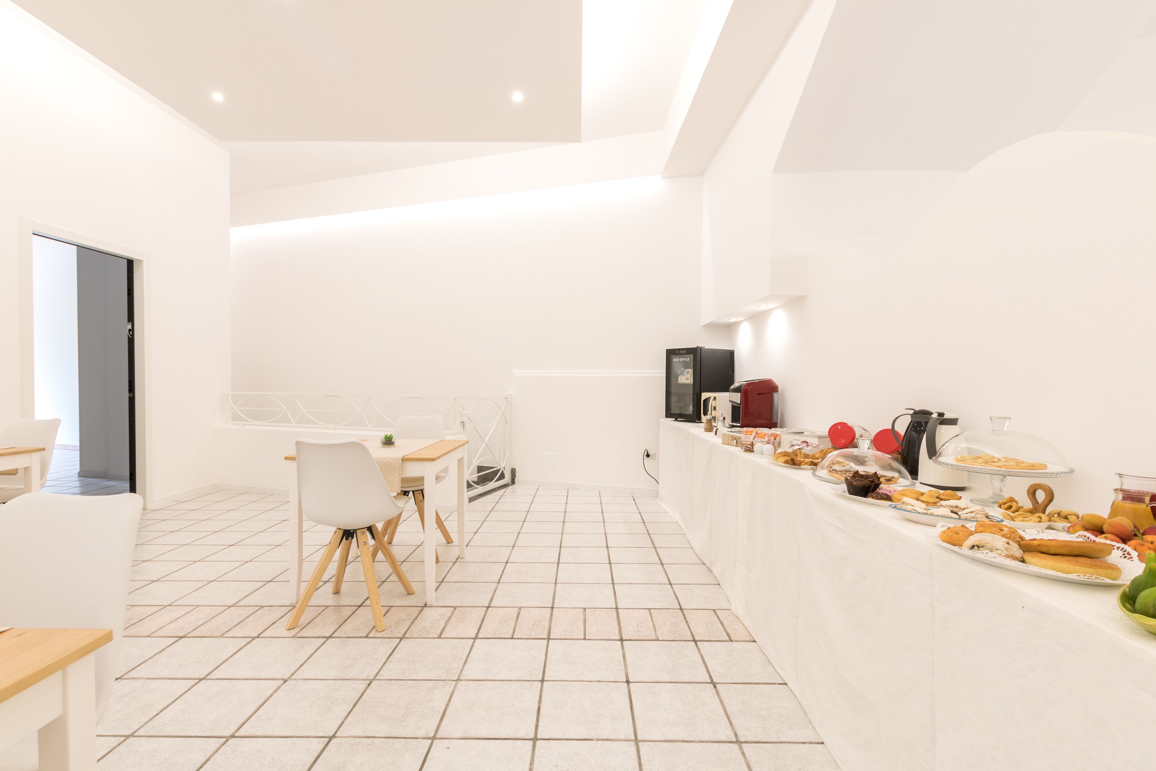 A bright, modern dining area with a long buffet table filled with various food items, a white dining table with chairs, and a clean, tiled floor.