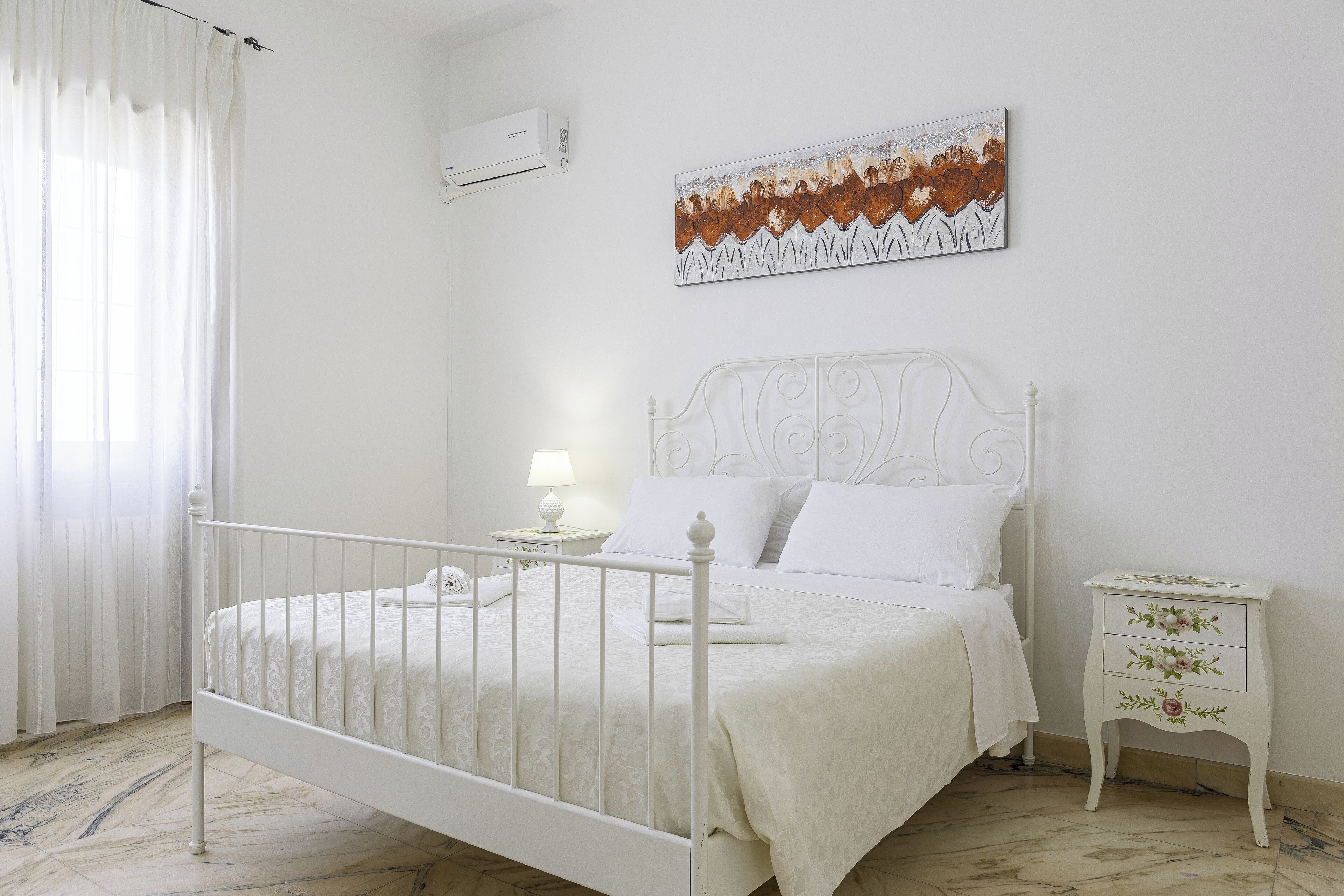 Un dormitorio blanco con una cama, una mesita de noche, aire acondicionado de pared y un cuadro.