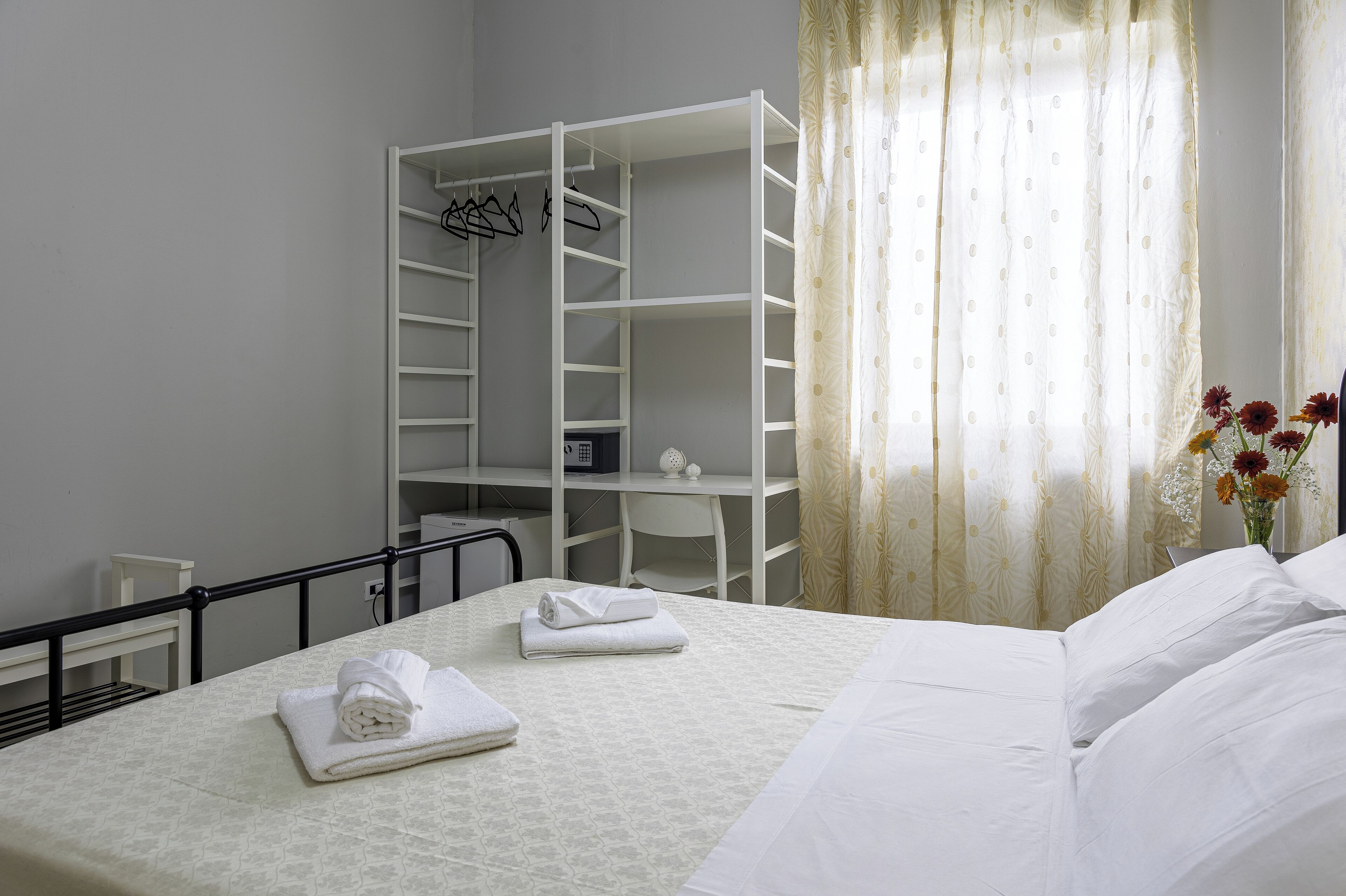 A neatly made bed with white linens, a white wardrobe with shelves, and a small table with a white cloth.