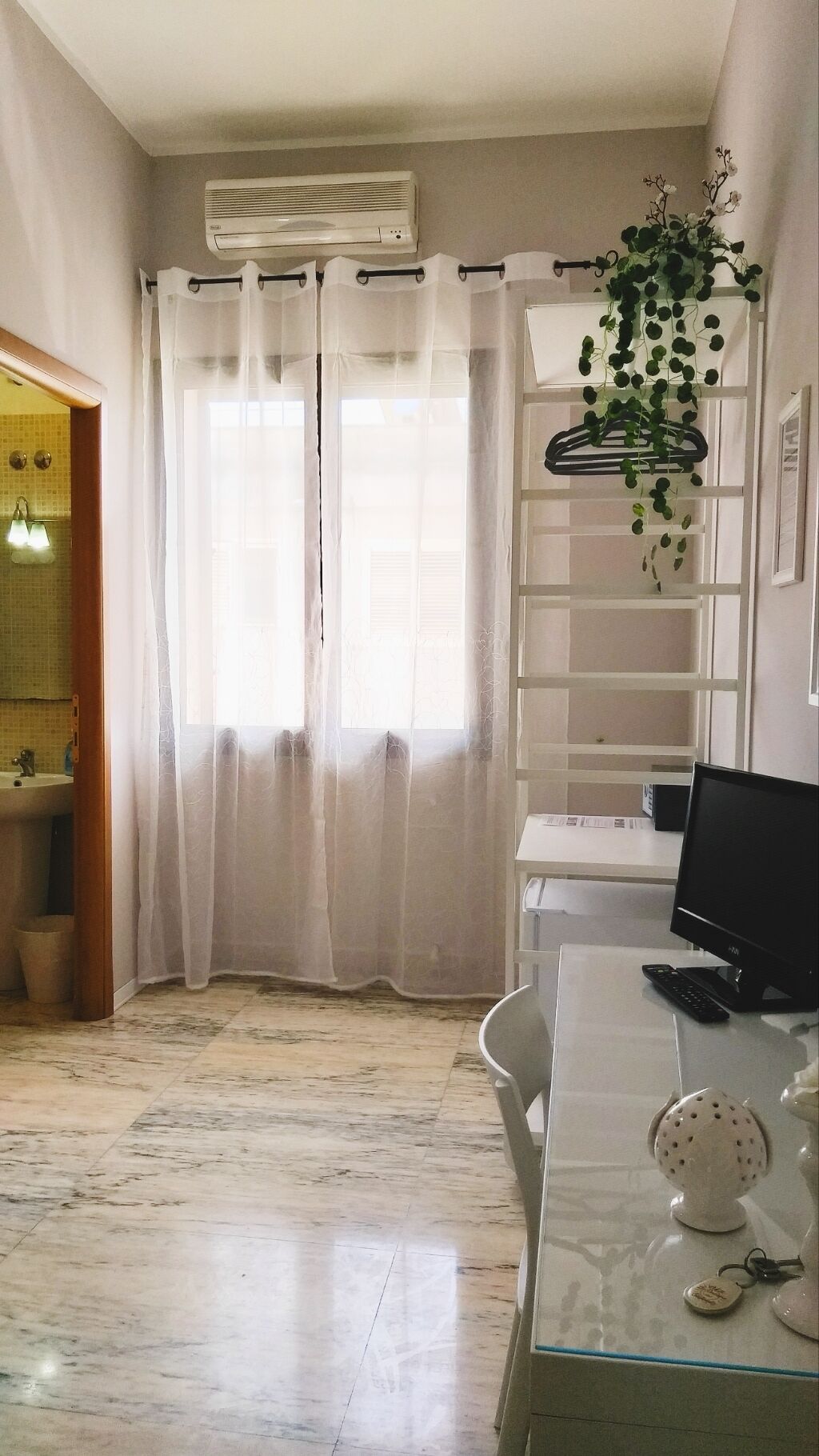 Ein Zimmer mit großem Fenster, Klimaanlage, Schreibtisch mit Computer, Stuhl und einer hängenden Pflanze.