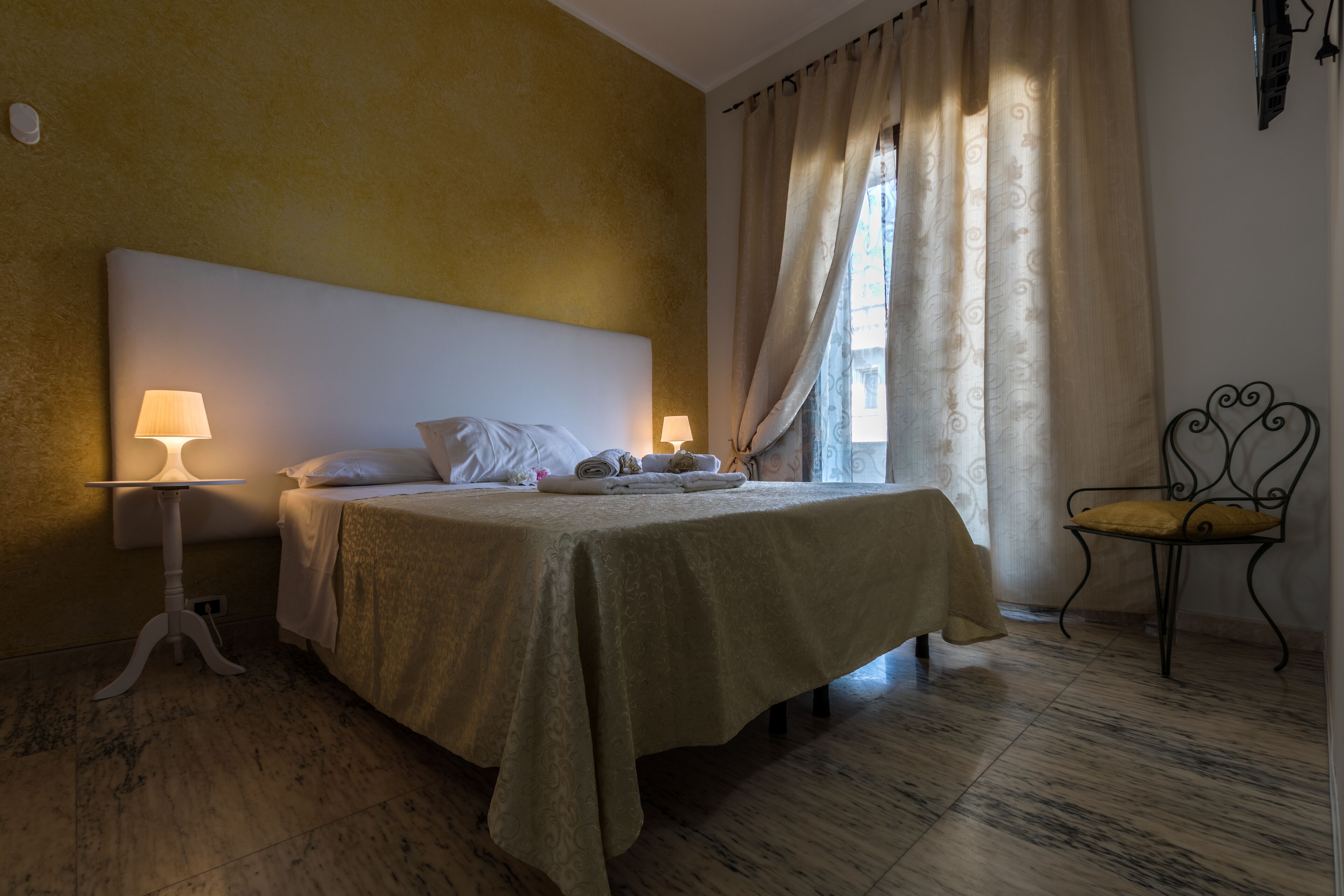 A neatly made bed with a headboard, two bedside lamps, a chair, and a window with curtains.