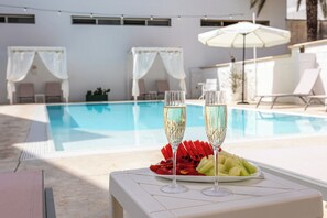 Escena junto a la piscina con dos copas de champán, una bandeja de frutas y una mesa blanca.